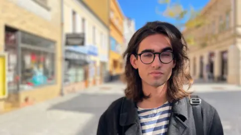 Onrej Kapralek is a dark -shoulder young man. He wears glasses with a black frame, shirt and striped jacket. He stands in a pedestrian area with stores on both sides. 