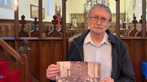 Kerry Devine/BBC David Edwards has short grey hair and a matching moustache he has glasses on and a blue fleece with a light blue shirt underneath it, he is holding an old black and white photo of the original concert from 1926 behind him are the choir stalls of the church and a stained glass window further back.