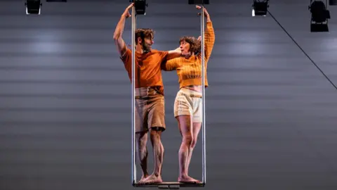 Ockhams Razor Charlotte and her partner stood on a trapeze in the air looking at one another and mirroring each others movement. They're both wearing shorts and orange tops.