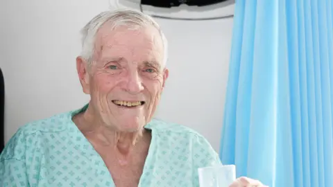 Cambridge University Hospitals NHS Foundation Trust Roger Jackson smiling at the camera while in a hospital. He has white hair with a side parting and is wearing blue-green scrubs with a pattern. He is holding a glass of water.