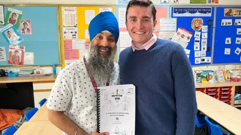 The two teachers stand in a classroom holding one of the packs 