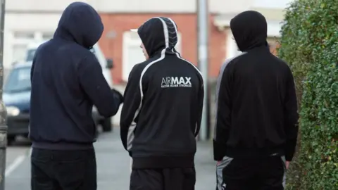 Three men, dressed in black hoodies and trousers, have their backs turned. They are walking in a neighbourhood. 