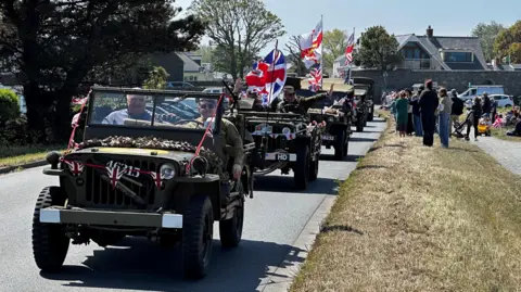 The image shows a procession of military vehicles decorated with flags, driving along a road by a grass bank with people watching.