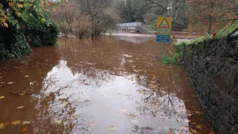 Severe flooding at Jubilee Bridge in Appleby. There is a road sign reading Ford, road liable to flooding. The water is a muddy brown colour with fallen leaves floating on the surface. 