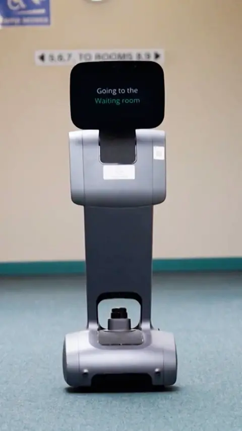 Robot in hallway of a medical centre with its screen reading "Going to the waiting room"