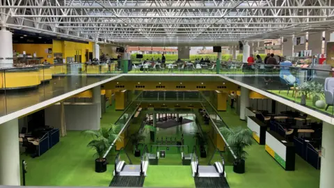 Matt Marvel/BBC The interior of a large, open-plan office building. Three floors are visible. The lower one has a modern entrance with pass barriers. Two escalators take people to the next floor, which has green flooring. The third level is a mezzanine one with yellow, green flooring, walls and desks. The ceiling has metal struts and glass above.