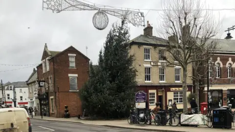 John Devine/BBC A 9m (30ft) Christmas tree which is leaning to the right in the Market Square, March. Shops are behind it and large decorations hang across the road in front of it