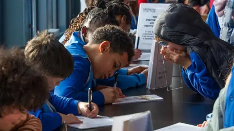 School pupils wearing blue uniform lean on a table inside a hall while they sign books. Another youngster waiting to pick up a book holds his hands over his face in excitement.