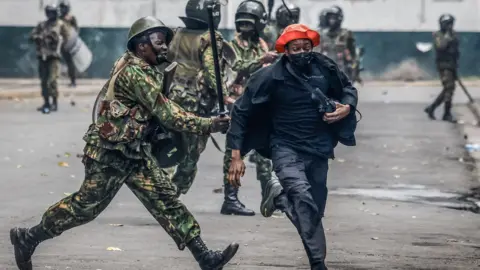 A Kenya police officers, in camouflage uniform, hits a fleeing protester, wearing a red hat, with a baton during a protest.