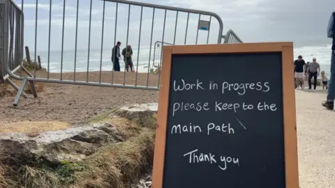 An area near a beach or coastal path. A metal barrier is set up to block access, and in the foreground, there's a blackboard sign with white text that reads: "Work in progress, please keep to the main path, Thank you"