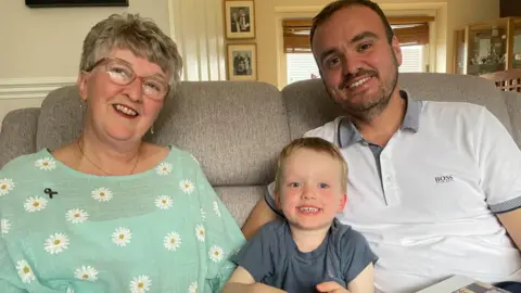 Simon Thake/BBC An older woman, a young boy and a middle-aged man pose together on the sofa smiling