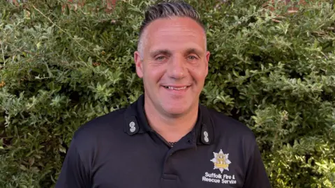 Mark Cowling is smiling and looking at the camera. He is wearing a shirt with the Suffolk Fire and Rescue Service badge and logo. 