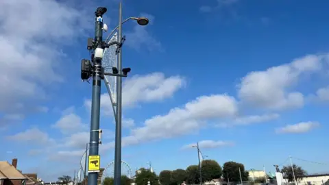 A sound camera with microphones and cameras, and aerials is mounted on a standard, next to which is a decorative street lamp. In the lower part of the image are trees and buildings on Great Yarmouth Sea Front. The sky is blue with several white clouds.