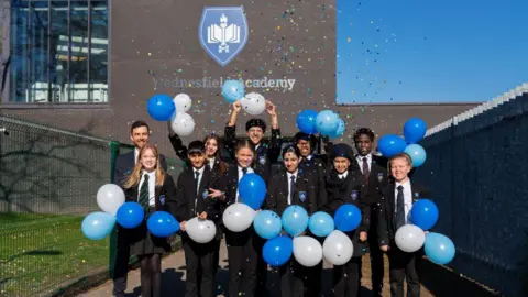 Ten children in dark school uniform with white shirts holding balloons alongside a man in a dark suit, all standing in front of a large brick building with a sign reading Wednesfield Academy on it