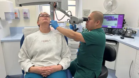 A man dressed in a green gown is looking through a microscope into a patient's ear