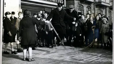 Hulton Archive/Getty Images A black and white photograph of two men, dressed in sailors' uniforms, skipping over a large rope. A crowd of children can be seen gathered around. 