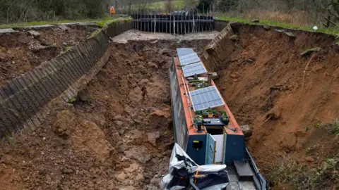 A long narrowboat with an orange roof and solar panels at the bottom of a deep muddy trench