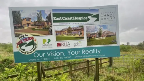 Andrew Turner/BBC A sign board showing the architects impression of the original plans for a hospice in Gorleston. It features a timber clad building with sweeping curved roof nad glazing panels. The logo of East Coast Hospice, the proposed name of Margaret Chadd House and the slogans: "Help to Raise the Roof of East Coast Hospice" and on the bottom banner, states: "Our Vision, Your Reality"
