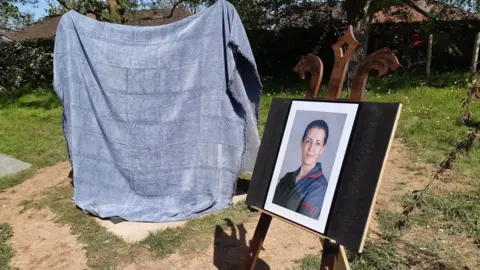 LDRS A photo of a nurse in uniform on an easel next to a monument draped in a grey blanket. It stands in a garden, with a bare patch of earth round it. The nurse wears a grey uniform with red piping around the sleeve and collar and has bown hair tied back.