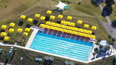 A computer-generated aerial image of an outdoor swimming pool with yellow sunshades that is surrounded by greenery, trees and pedestrian walkways.