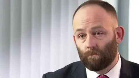 A man with a beard wearing a suit looks on forlornly while sitting indoors.
