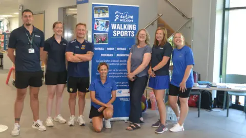 Wiltshire Council Seven people stand around a vertical banner which reads "Active Wiltshire Walking Sports". They are inside a leisure centre foyer and are all wearing activewear, with most of them in shorts and dark blue T-shirts.