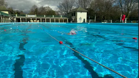 Emma Baugh/BBC An outdoor lido swimming pool. At least one person is swimming lengths. There are ropes dividing up the lanes.