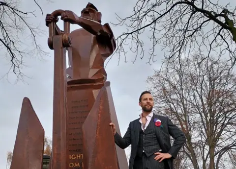 Sandwell Council The camera is looking up at the monument, which Luke Perry is standing next to, and there are trees in the background.