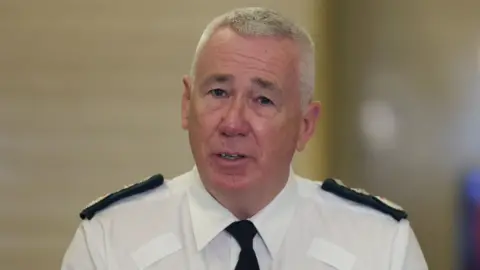 Liam McBurney/PA Wire PSNI Chief Constable Jon Boutcher, a man with short, grey hair, speaks to the media during a previous press event. He is wearing a white uniform shirt, a dark-coloured tie and dark-coloured epaulettes with gold details. 