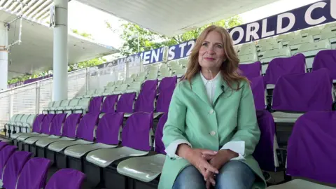 A person with shoulder-length, wavy, reddish-brown hair sits in a covered cricket stadium stand, wearing a light green coat over a white collared shirt and blue jeans. The individual is seated with hands clasped in their lap. Rows of empty pale-green seats surround the person, many draped with purple seat covers. In the background, signage reads “Women’s T20 World Cup,” and trees are visible beyond the stadium structure.
