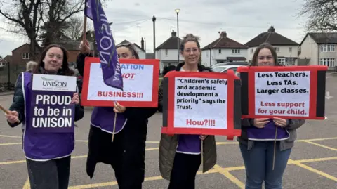 BBC / Charlotte Henderson The photo shows four women standing in front of the camera. They're all wearing purple tabards, and are holding various signs and purple flags which read UNISON on them. 