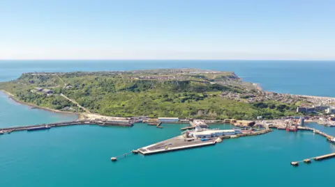 The Isle of Portland in birds-eye view from the sea, showing the port and greenery of the island, surrounded by the sea. 