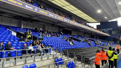 Connor Bennett/BBC A close up view of one of the stands at the stadium that has two levels. The chairs are blue, and in long rows which span across the whole picture. People are sat staring towards the right of the image where the boxing ring is.