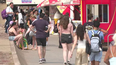 The image shows a busy seaside promenade on a warm day, with large numbers of people walking and gathering outside food kiosks and takeaway stands. A bright red snack kiosk dominates the right side of the picture, while ice cream signs, parasols and groups of people carrying bags and backpacks suggest a popular tourist area during the summer season.
