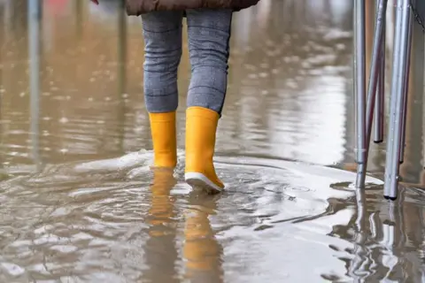 Getty Images A person wearing yellow wellies walks through a flooded street caused by rain water.