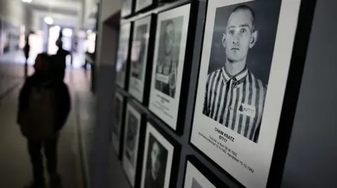 Getty Images Black and white portraits of prisoners line a wall at the former Nazi German concentration camp Auschwitz-Brikenau. People visiting the site are walking down the corridor.