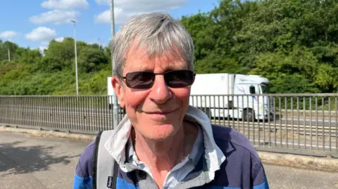 John Devine/BBC Harry Machin has short grey hair swept across his forehead. He has dark glasses on and a light blue and dark blue hooped rugby style shirt on, with a white shirt underneath. Behind him are metal railings and a busy roadway, with a white lorry passing.