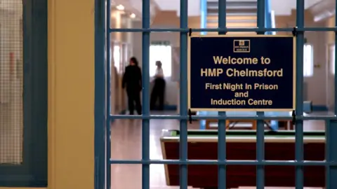 An image of a prison sign that reads Welcome to HMP Chelmsford. There are two people standing in the frame and there are also stairs in the background. 
