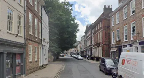A road with two and three storey buildings on either side. Cars and vans parked along the right-hand side of the road. A large tree on the left side of the road.