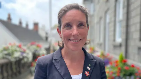 Deputy Lindsay de Sausmarez, a woman with brown hair smiling at the camera. She's wearing a blue jacket and a white blouse. On her lapel are three pin badges.