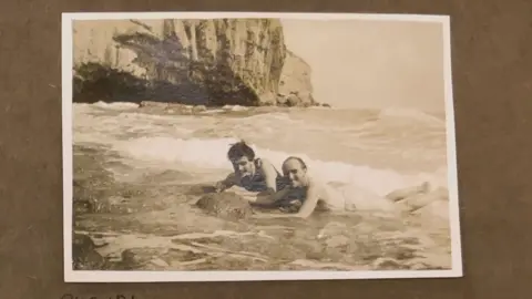 Dorset Museum A black and white photograph of David Brynley and Norman Notley laying on their front in the shallows of the sea in swimming trunks. Behind them is a large cliff.