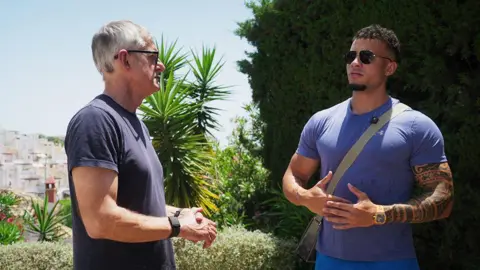 Netflix Louis Theroux speaks to HS TikkyTokky in a sun-kissed corner of Marbella overlooking the resort town. Both men wear t-shirts and clasp their hands together while locked in conversation. HS TikkyTokky wears aviator sunglasses and his muscular arms are visible.