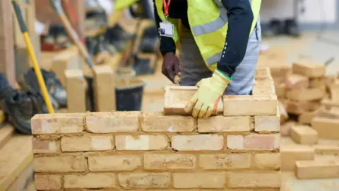 A man in a yellow reflective jacket with yellow gloves on is laying a light brown brick on a small wall. There are other bricks behind, buckets and tools behind him.