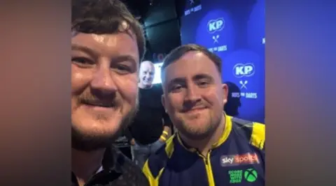 Samuel Caswell Samuel Caswell is on the left, with short dark hair and a beard. On the right is Luke Littler, who can be seen in his professional darts blue and yellow shirt. He also has short dark hair and a beard. They are both smiling and are standing in front of a blue background reading KP Nuts for Darts. 