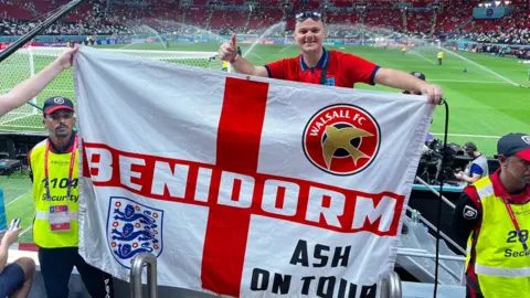 Ashely Price holding a St George’s flag with Walsall FC and England emblems at a football stadium, standing by the pitch before a match as sprinklers water the field and crowds fill the stands
