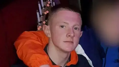 Jack McKenzie, a young man with short light brown hair and wearing a black top, looks at the camera - the arm of a friend, who we don't see, wrapped around his shoulder
