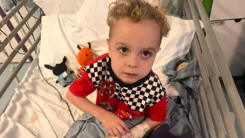 Family handout Kacey is pictured looking up from a hospital bed. The picture is taken from above the bed. There are two cuddly toys on the bed behind him, both characters from the CBBC show Bing. Kacey is wearing Lightning McQueen pyjamas which are red, white and black. He has a tube in his hand.