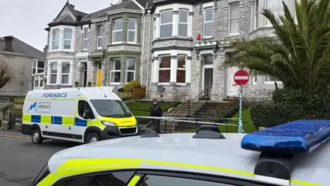 A row of stone built terraced houses with two police vehicles in front of them and a police officer standing outside.