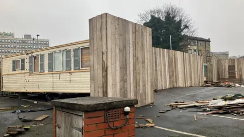 A white caravan unit resembling a mobile home with boarded-up windows sits on a car park. Around the side is wooden panelling partly obscuring it from view.