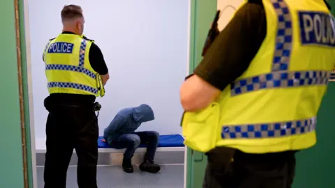 File photo of two police officers with someone wearing a hoodie and jeans sitting in a police custody cell.